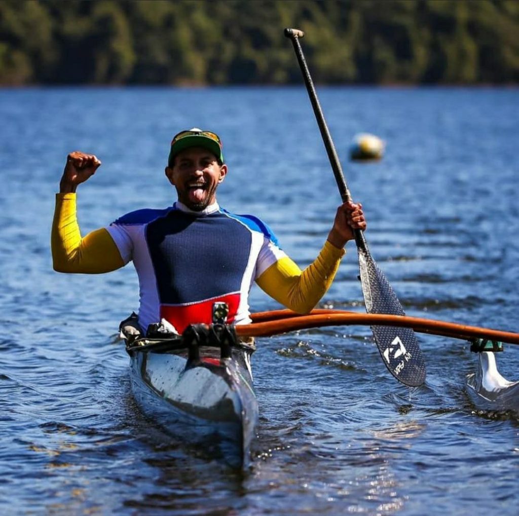 Homem branco, de boné, em canoa no rio. Braços erguidos em comemoração. Mão direita segura um remo levantado.