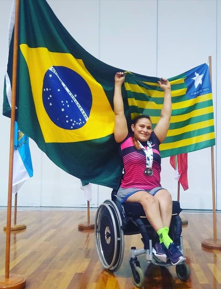 Mulher em cadeira de rodas com os braços erguidos em comemoração. Ao fundo estão hasteadas as bandeiras do Brasil e do Piauí 